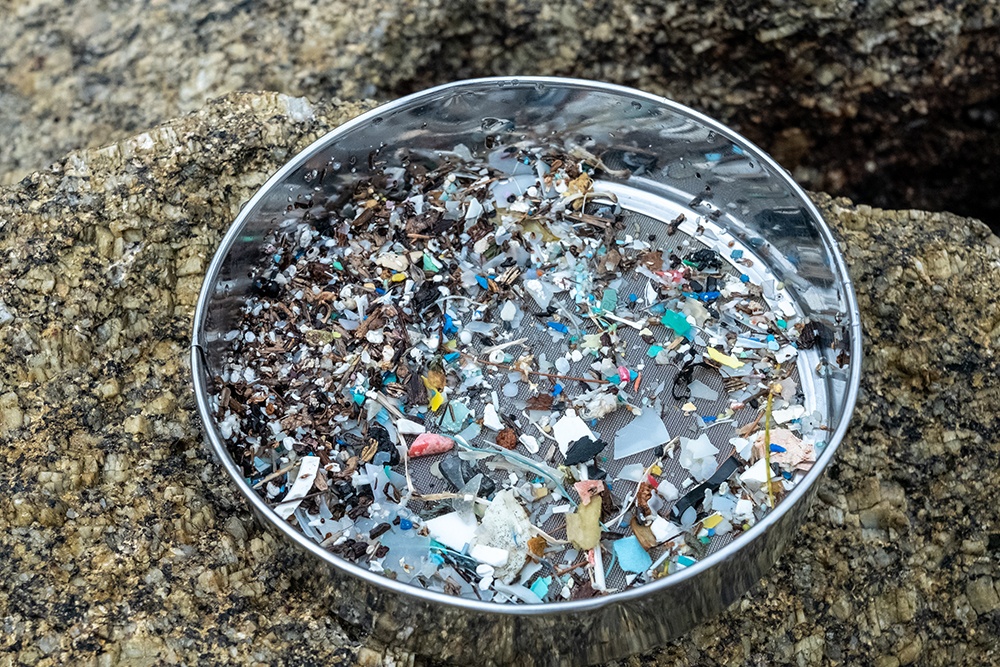 A photo showing a sieve containing colourful microplastics on a bed of sand